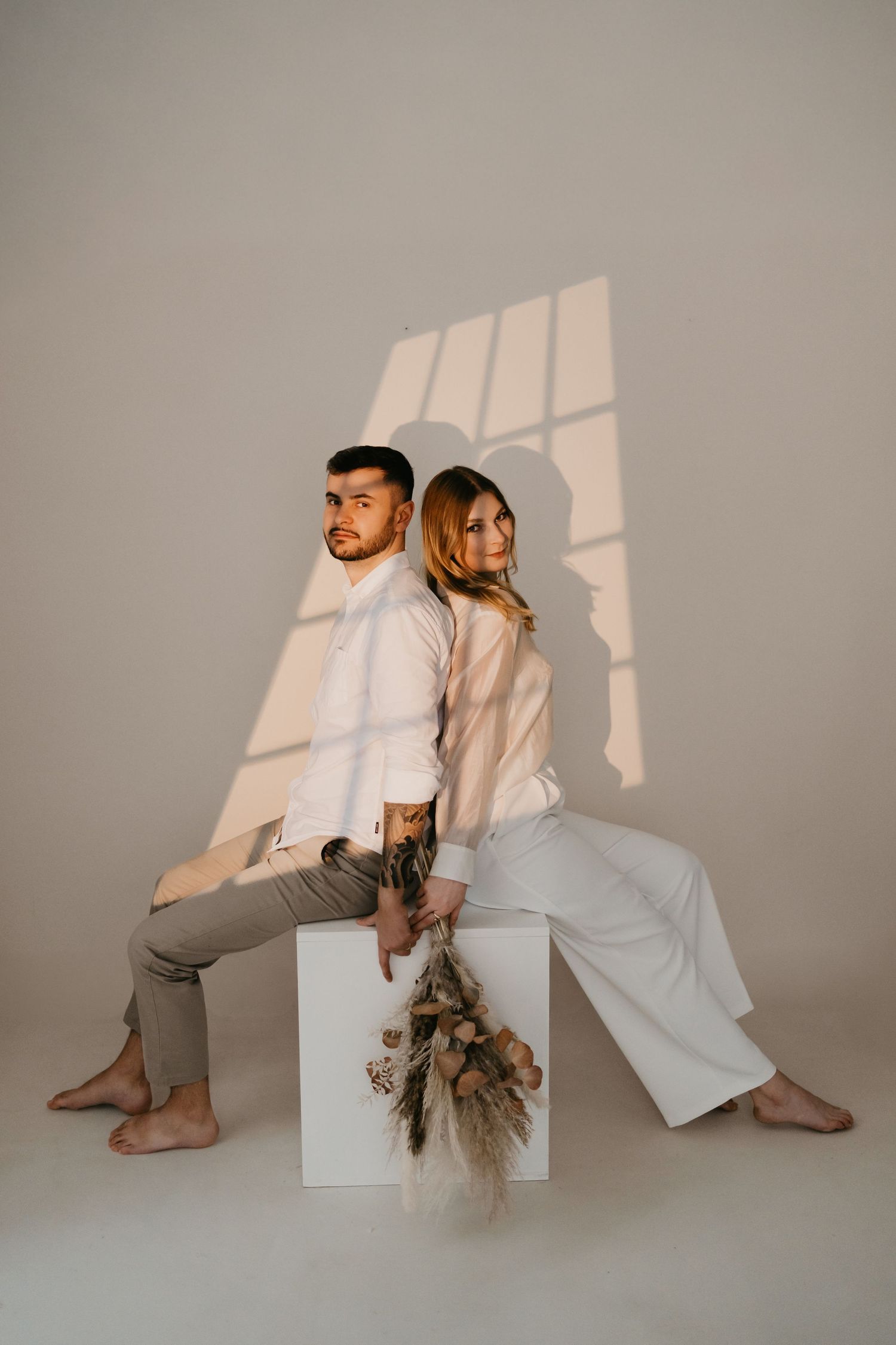Couple in neutral-toned outfits sit back-to-back on white pedestal with dried floral arrangement in minimalist setting.