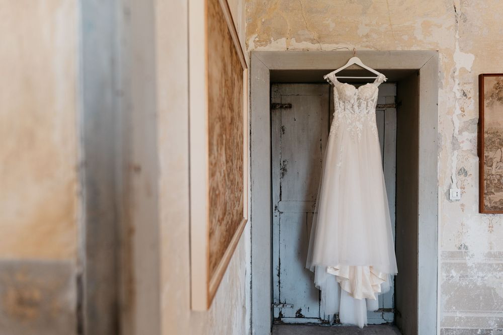 A white lace wedding dress hangs in a rustic doorway against textured walls.
