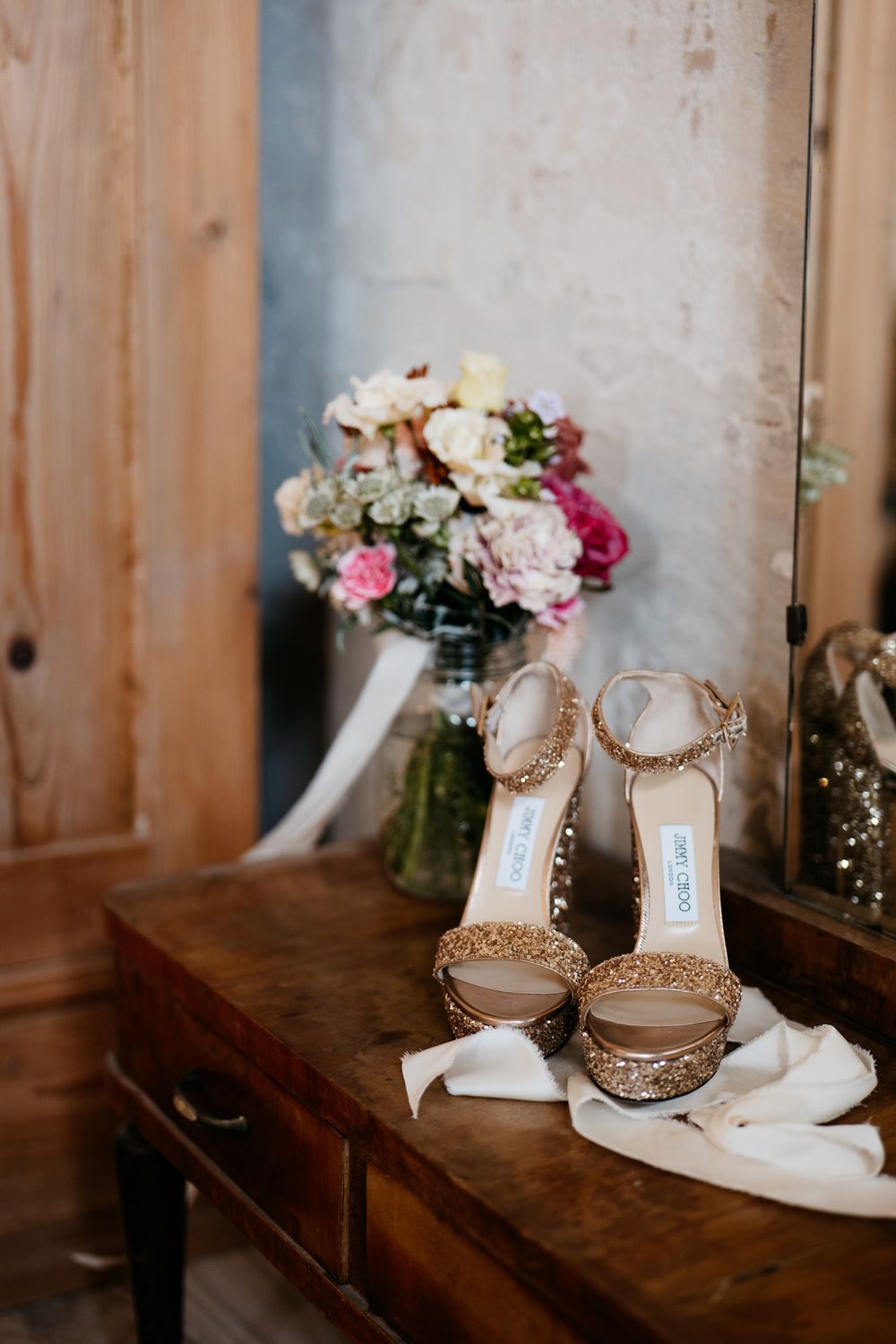Sparkly bridal shoes next to pink and white flower bouquet on wooden table.