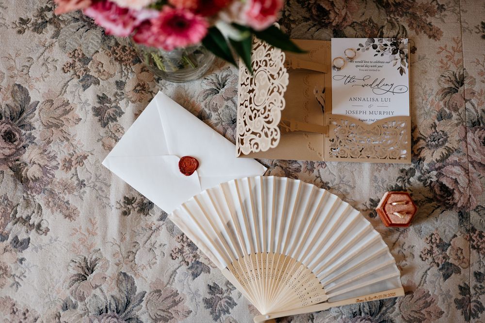 Wedding details arranged on vintage floral wallpaper including a white paper fan and pink flowers.
