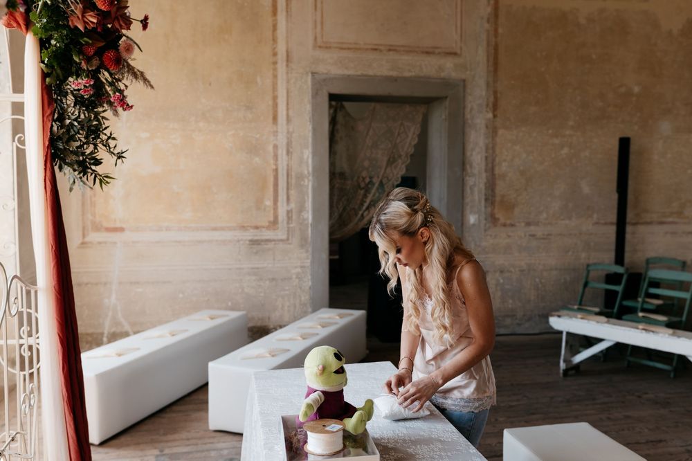 A bride in a flowing white gown sits on a bench in an elegant stone-walled room with red floral accents.