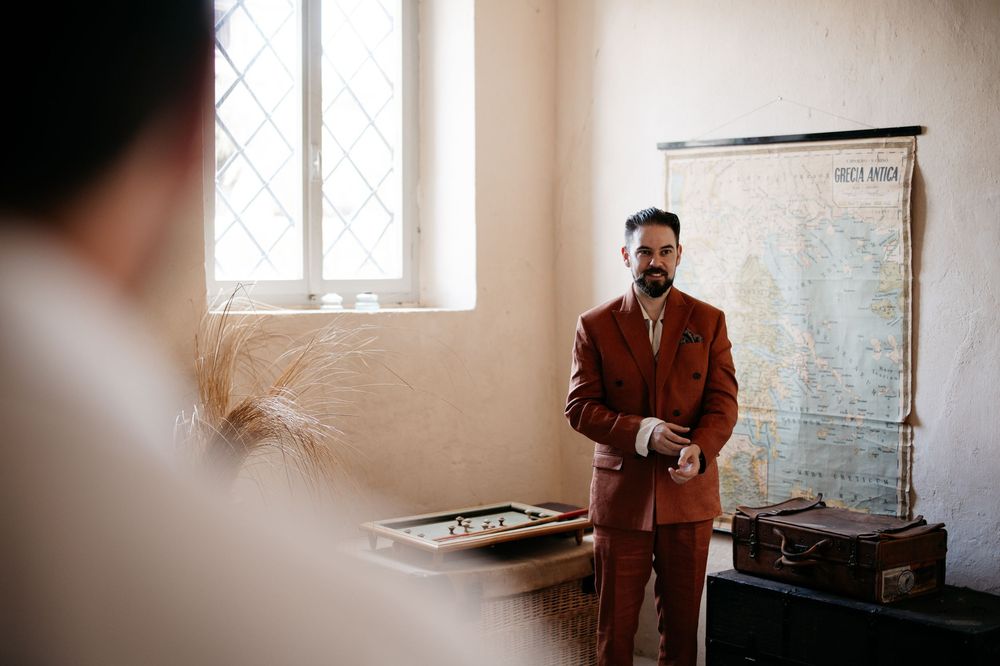 A person in a brown suit stands in a rustic room with distressed walls and natural window light.