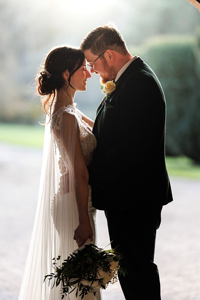 A romantic wedding couple shares an intimate moment outdoors with a flowing veil and dark suit silhouetted against nature.