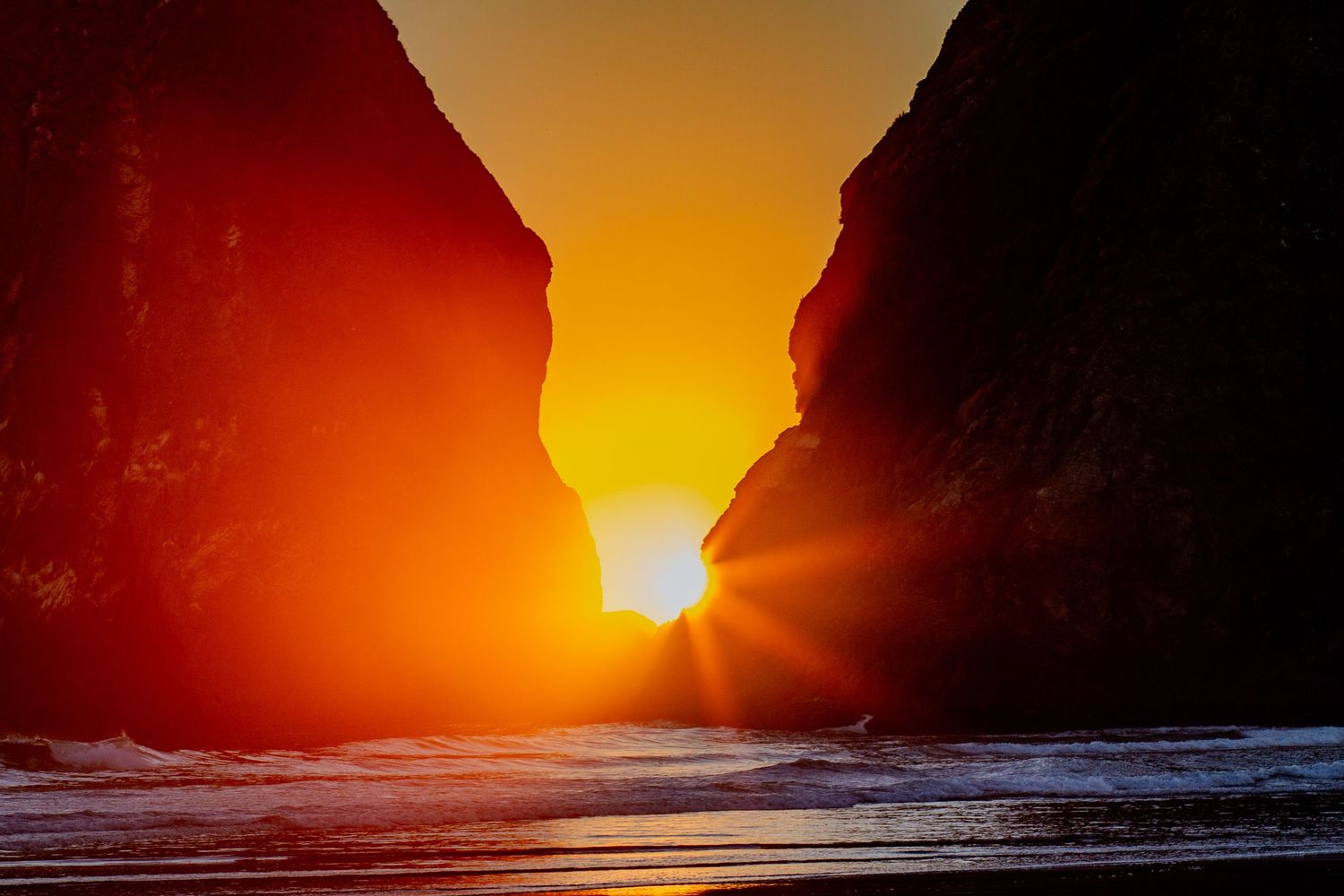 Dramatic orange sunset glows through a rocky coastal arch formation as waves crash against the shore.