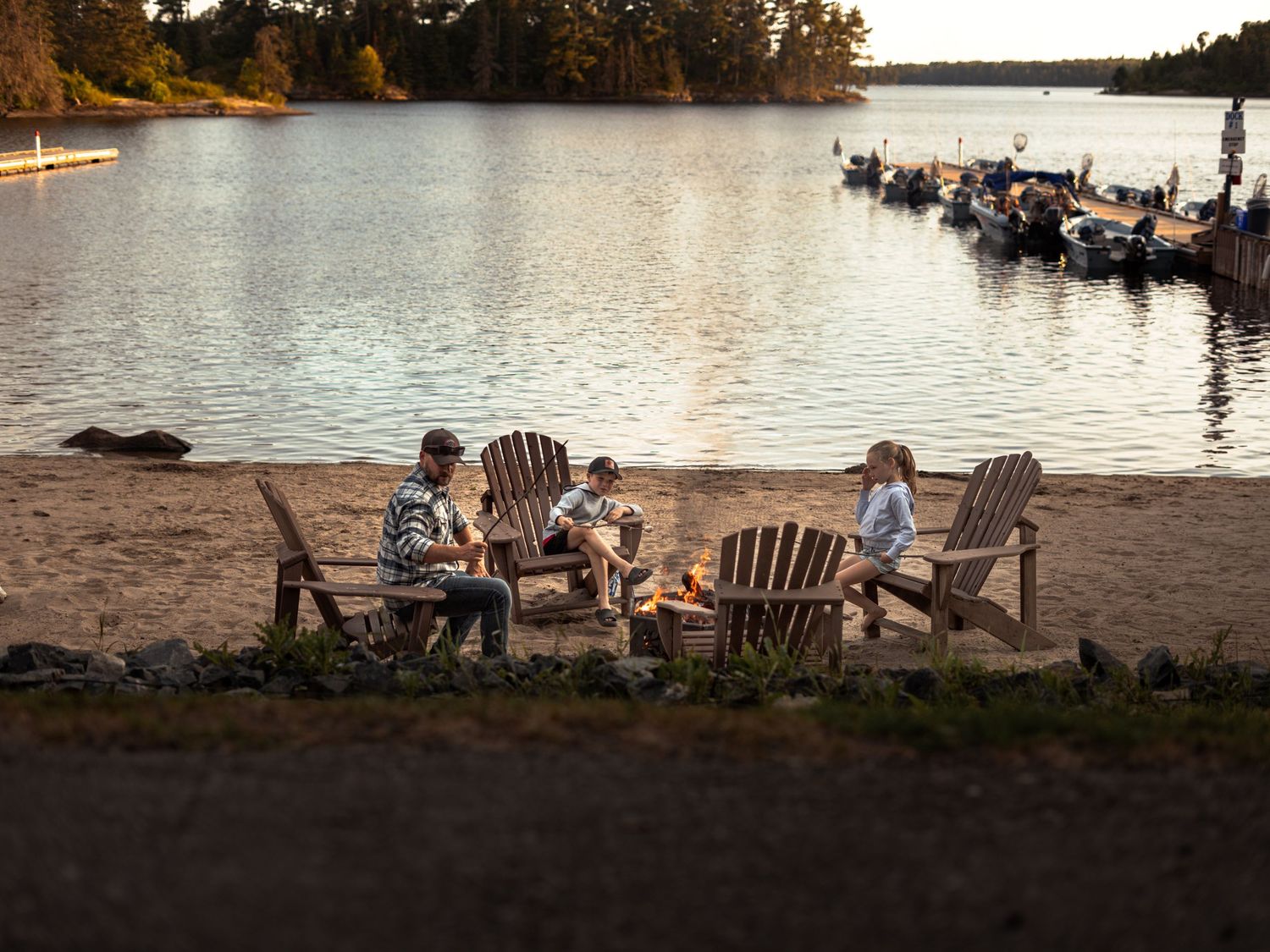 People relaxing in Adirondack chairs on a beach at sunset near a lake.