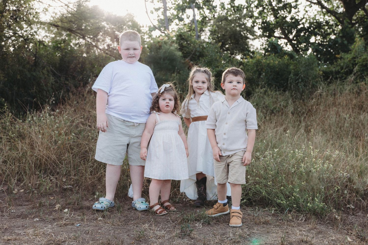Children in coordinated white outfits sitting together in a rustic outdoor setting.