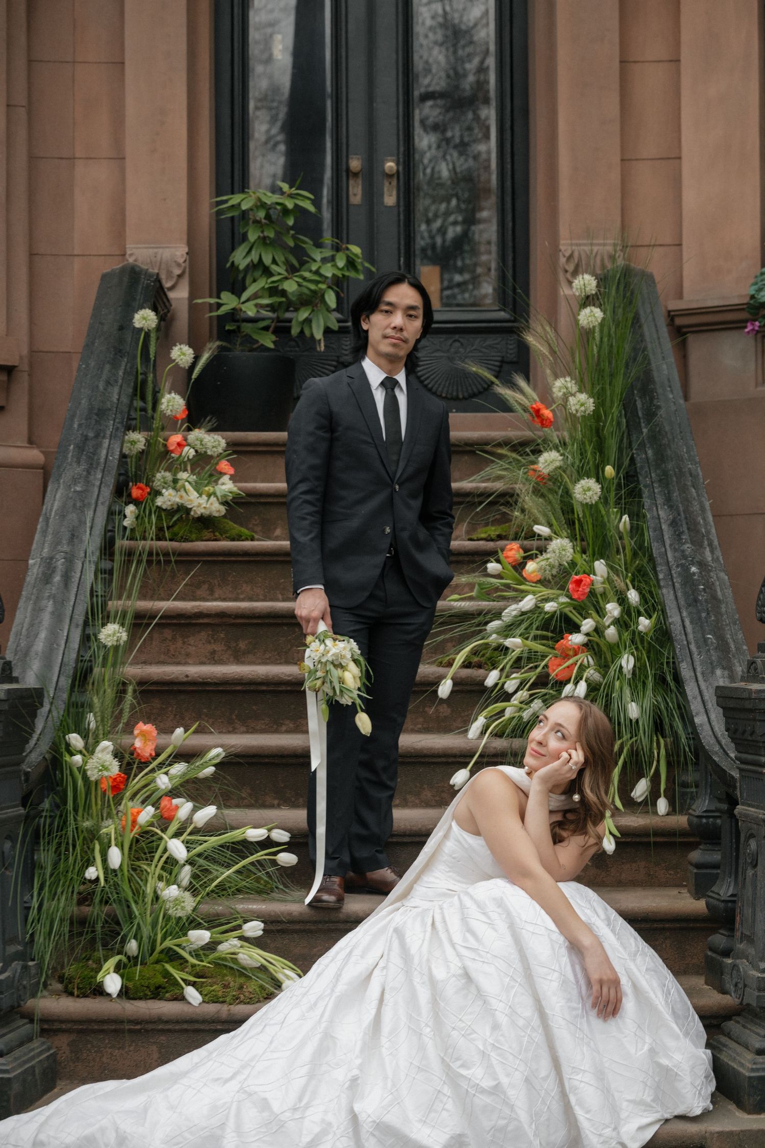 Bride in elegant white gown poses dramatically on stone steps decorated with floral arrangements.