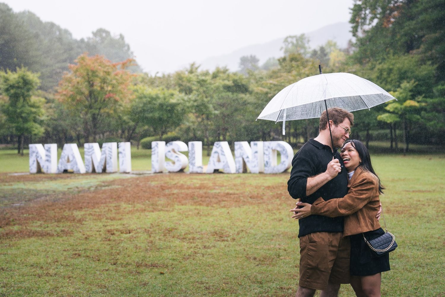 A romantic couple shares an intimate moment under an umbrella in a misty park setting during autumn.