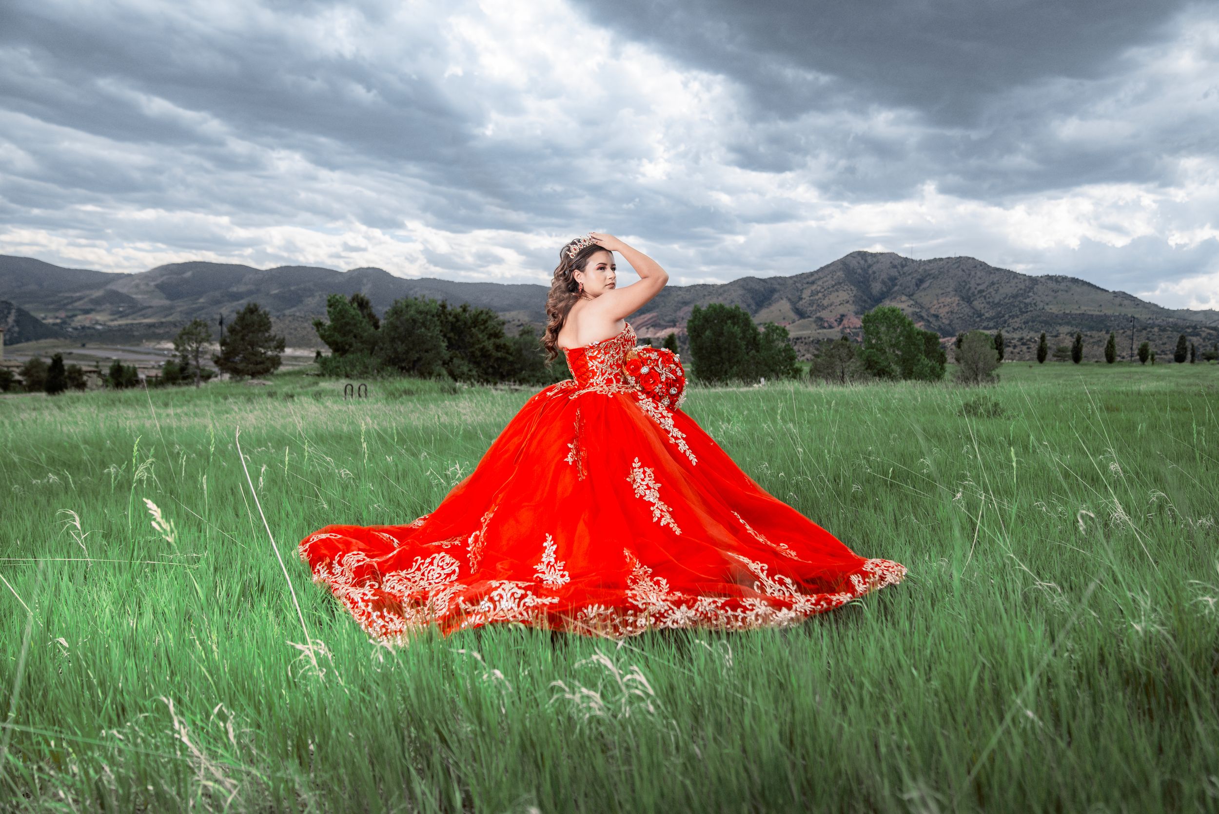 A figure in a flowing red gown poses dramatically in a green meadow with dramatic mountains and stormy skies in the background.