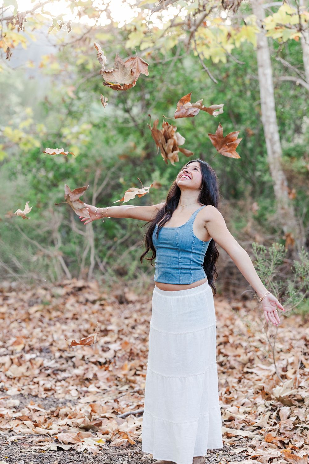 A person in a blue top and white skirt dances joyfully among falling autumn leaves in a wooded setting.