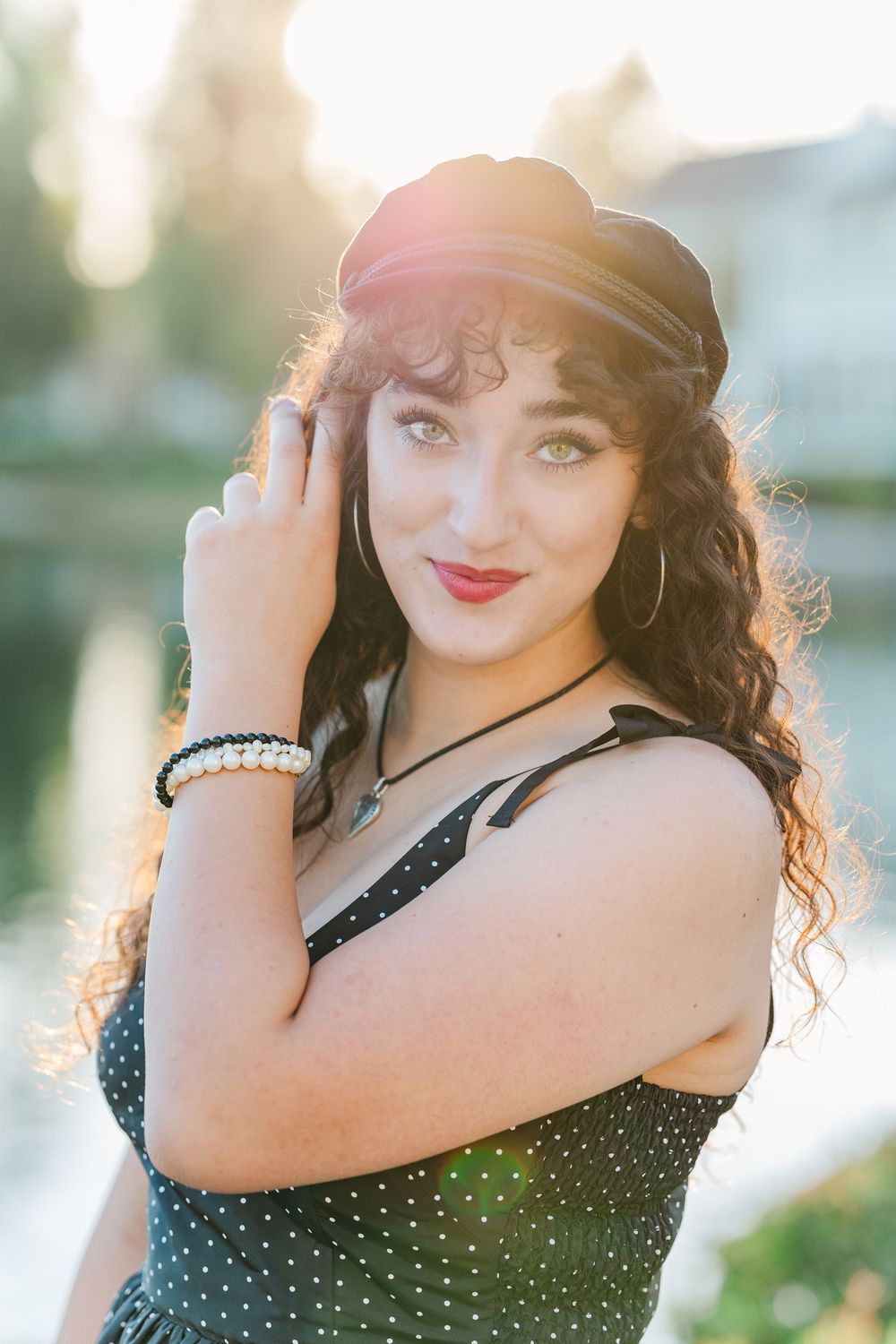 A person in a black polka dot dress and pearl bracelet poses outdoors in soft sunlight.