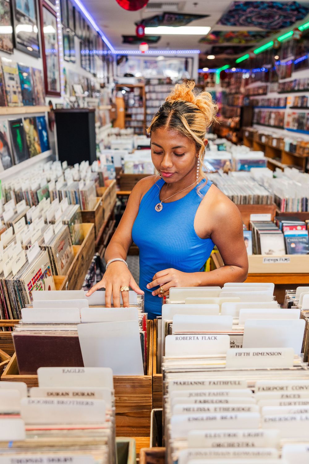 A customer in a blue sleeveless top browses through vinyl records at a music store with colorful lighting overhead.