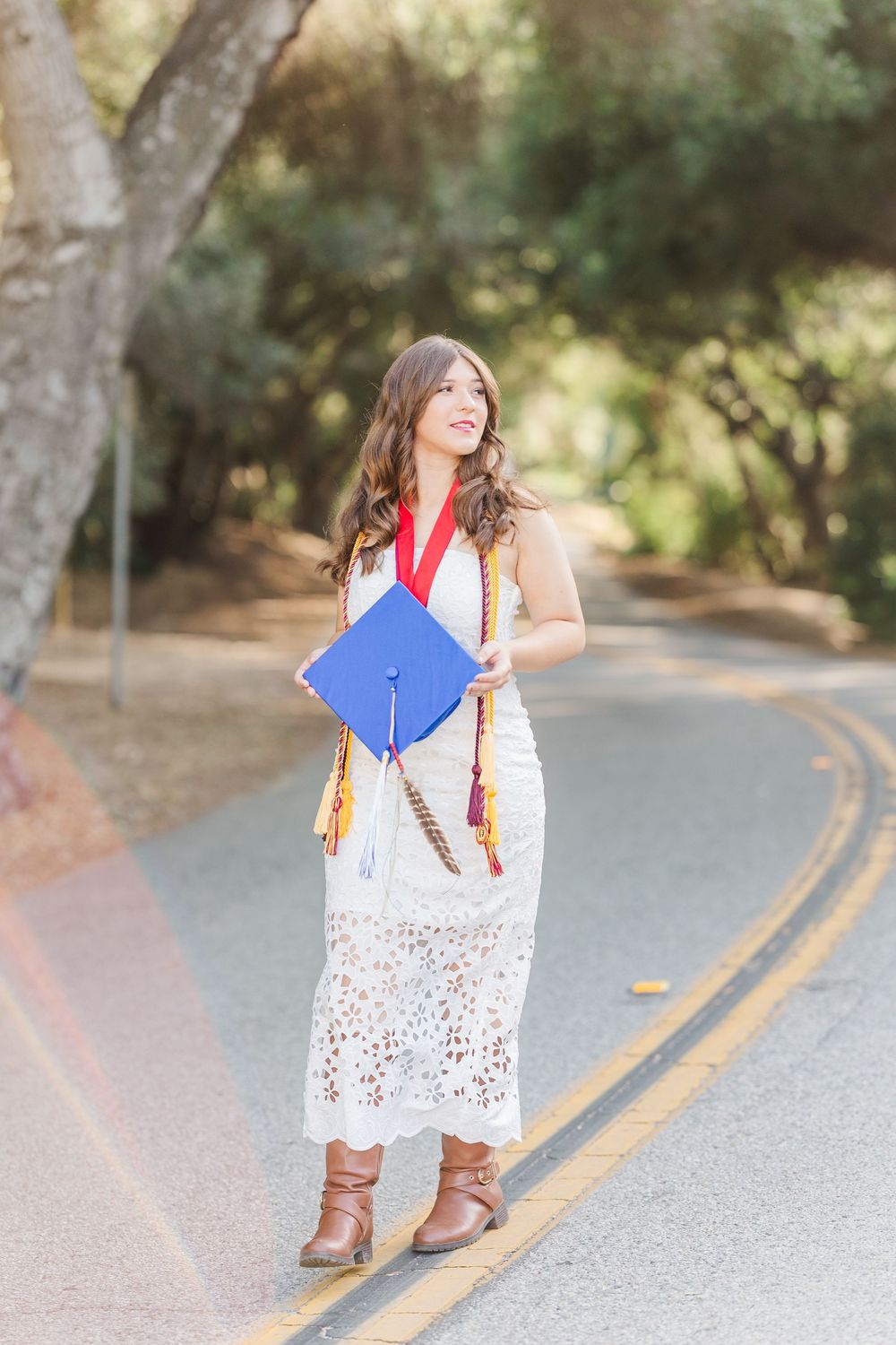 Graduate in white lace dress and red graduation sash poses on curved road with diploma in hand.