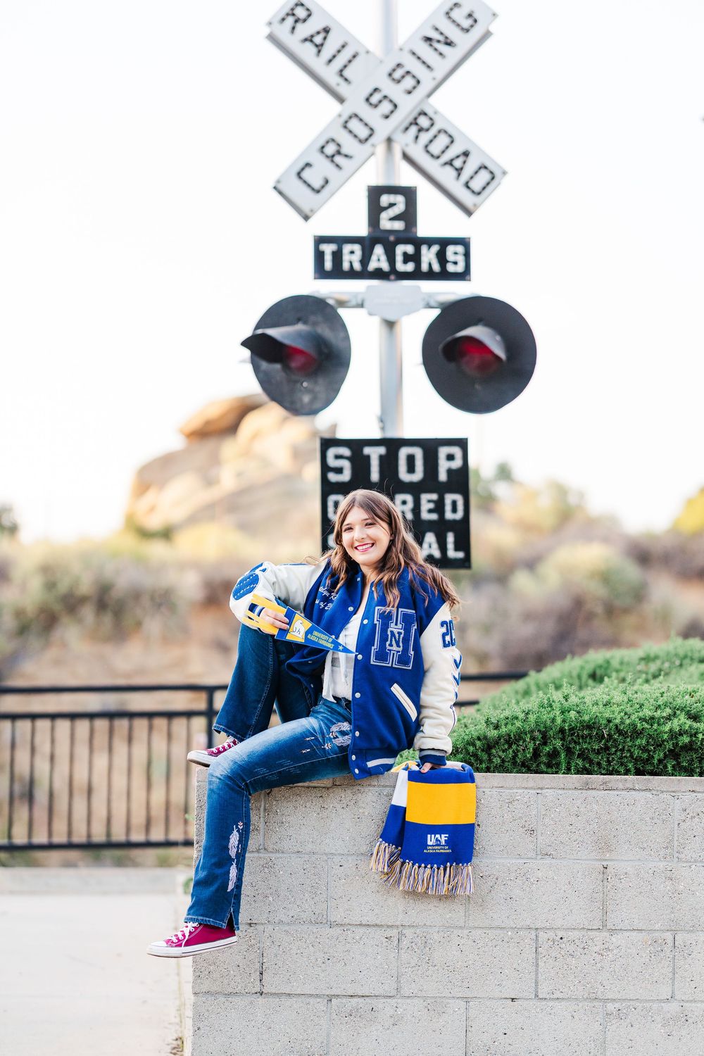 A person in a baseball cap and scarf sits on a concrete wall near railroad crossing signs in a casual outdoor setting.
