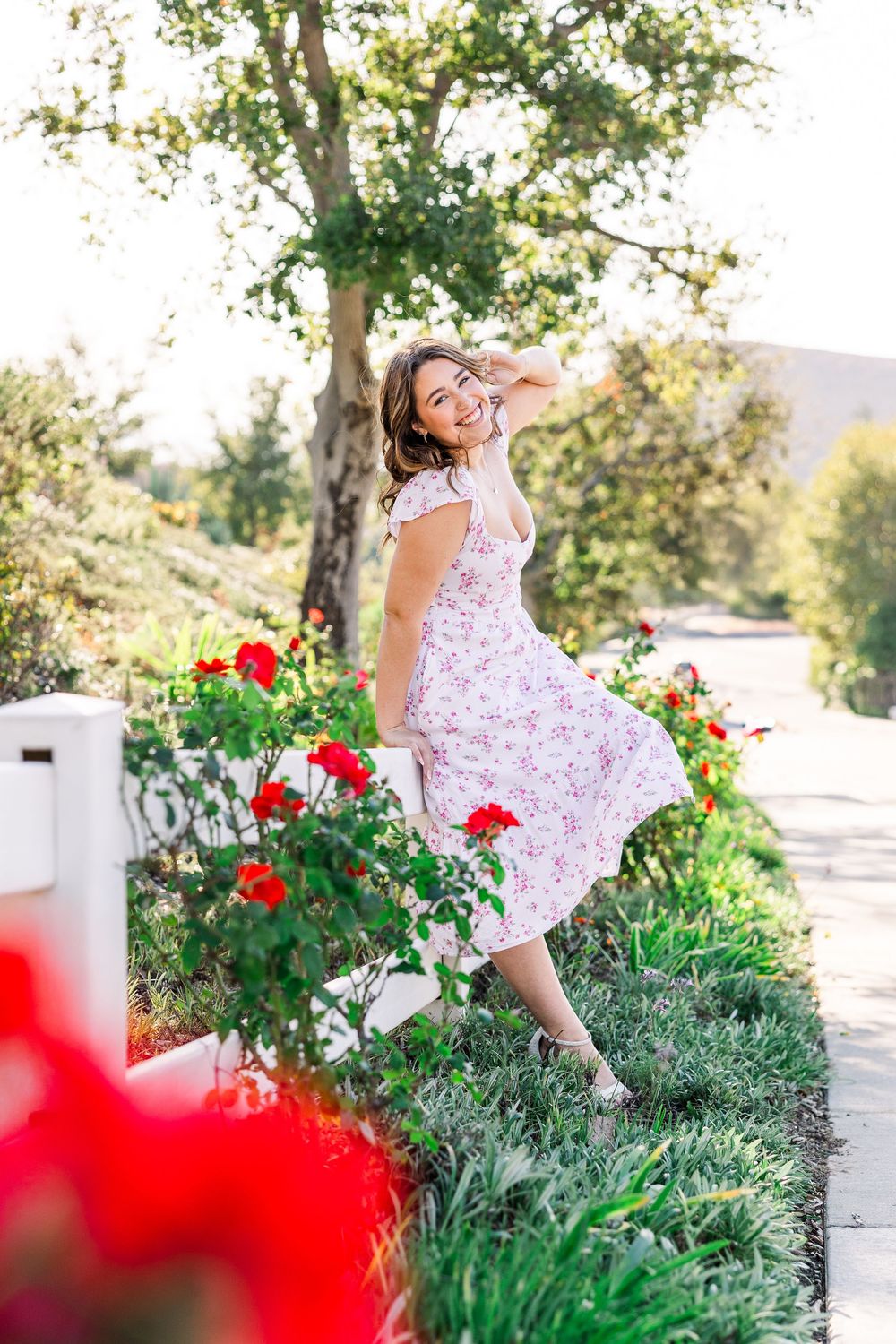 A woman in a floral dress twirls joyfully among vibrant red flowers and greenery on a sunny garden path.