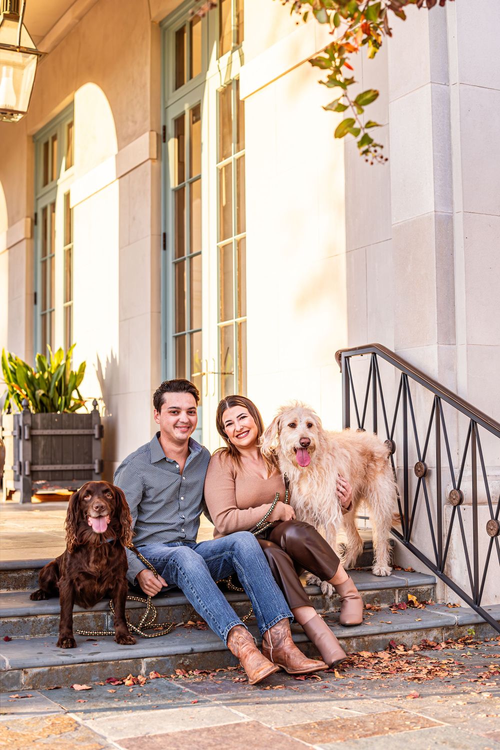 Couple sits on outdoor steps with their two dogs in front of elegant architecture.