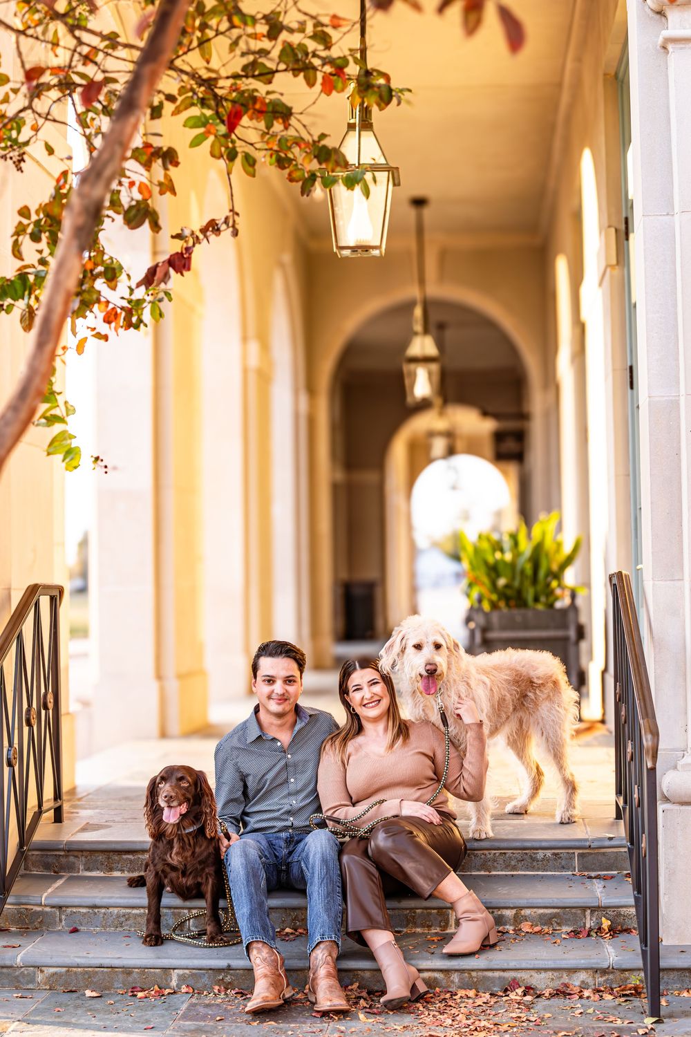 Couple and their two dogs pose casually on steps of elegant building with arched walkway.