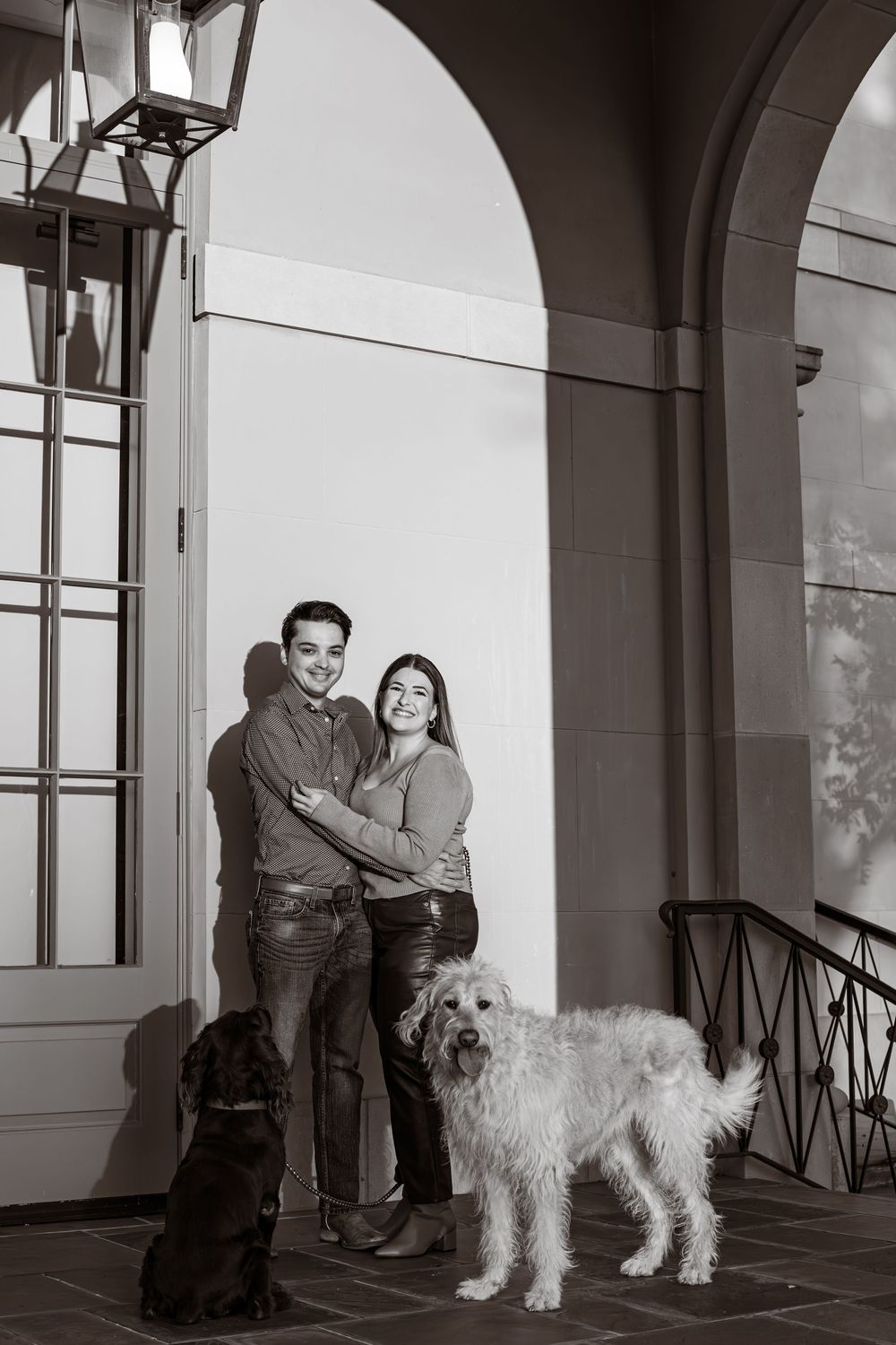 Black and white portrait of couple with their two dogs in front of arched doorway.