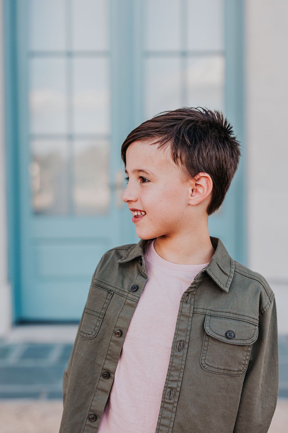 Young person in olive jacket smiling against blue door backdrop.