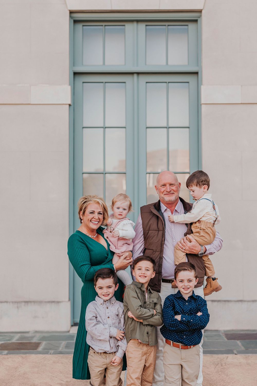 Multi-generational family group poses together in front of a tall blue-framed window wearing coordinated casual attire.
