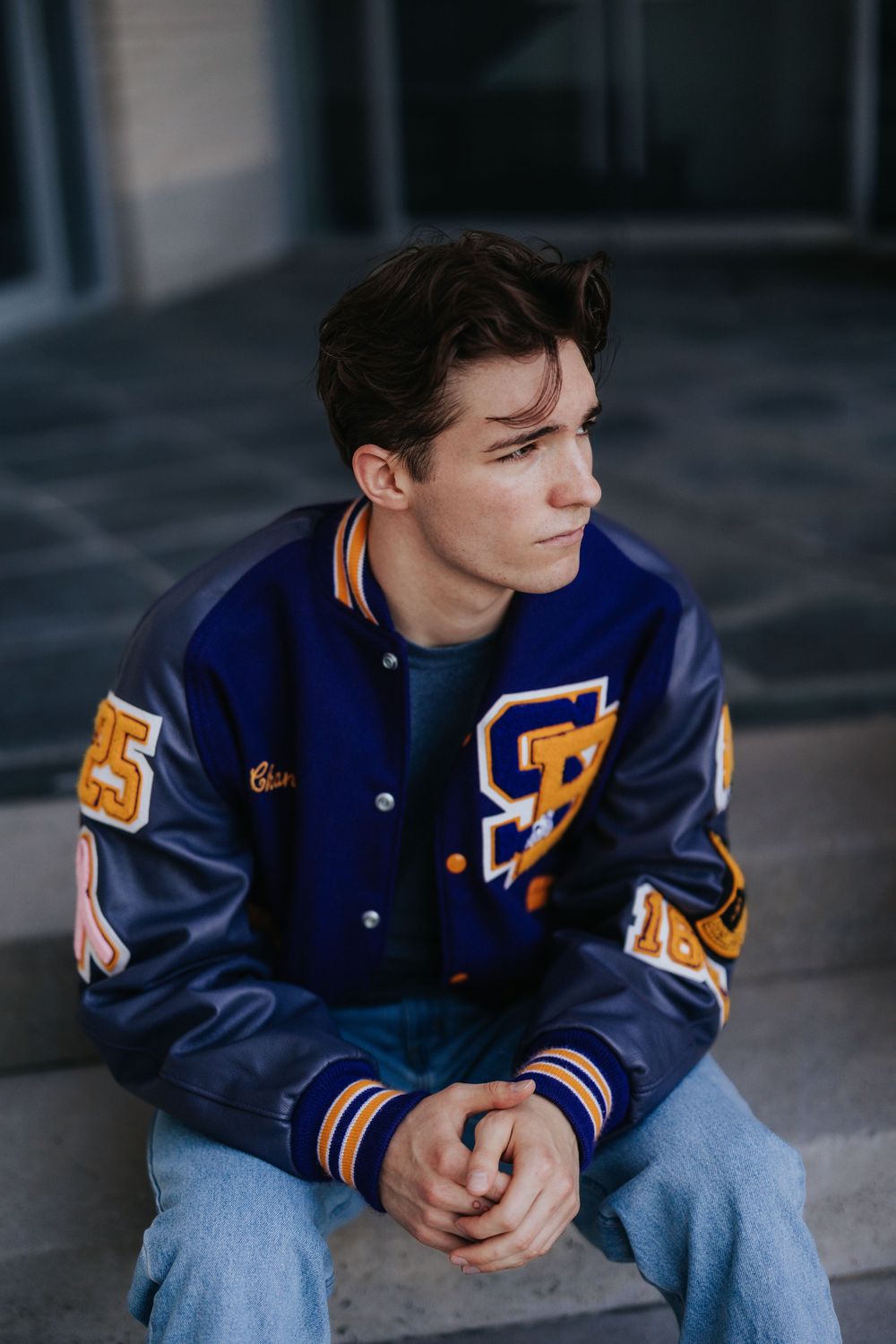 Person in navy letterman jacket sitting thoughtfully against dark background.