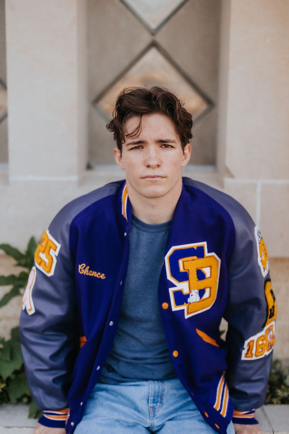 Person in varsity jacket and jeans sitting against geometric architectural background.