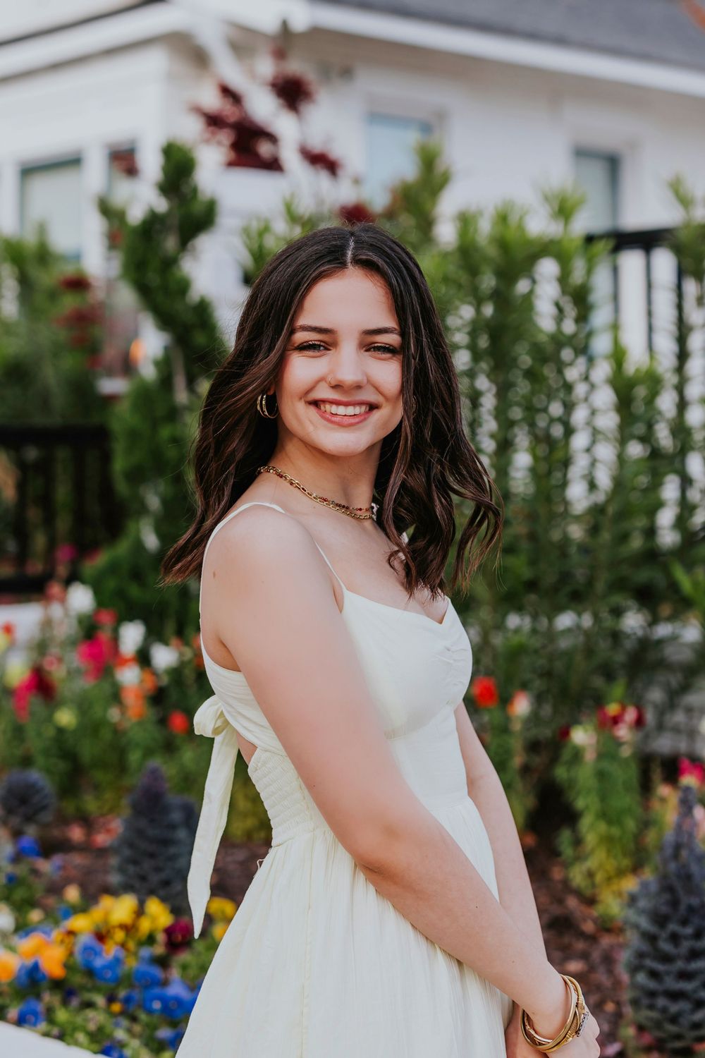 Person in white sundress poses among colorful garden flowers.