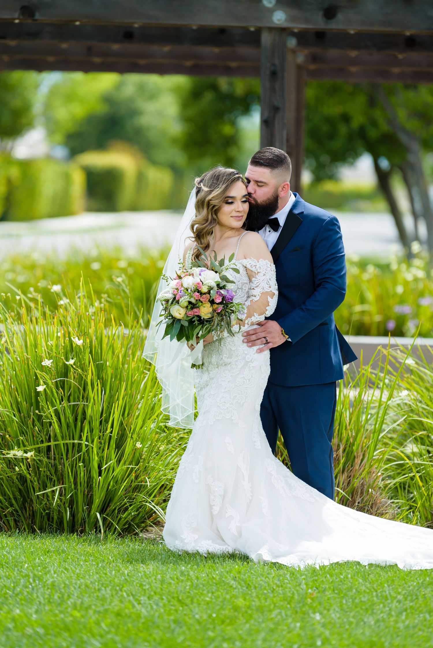 A romantic wedding couple embraces in a lush green garden setting with tall ornamental grasses and natural lighting.