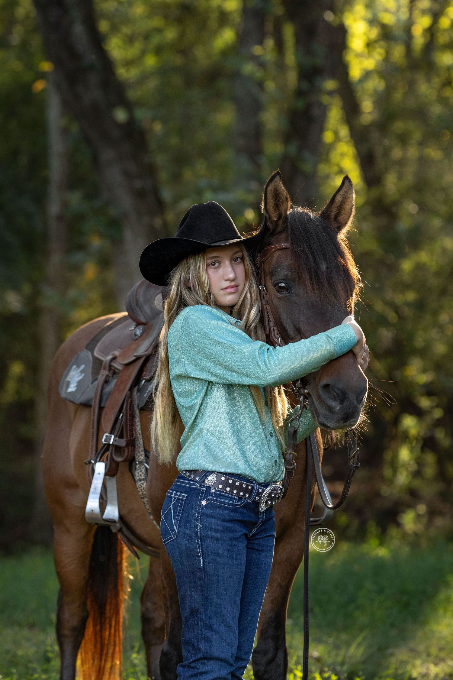 A rider in a turquoise sweater and cowboy hat embraces a brown horse in a sunlit forest setting.