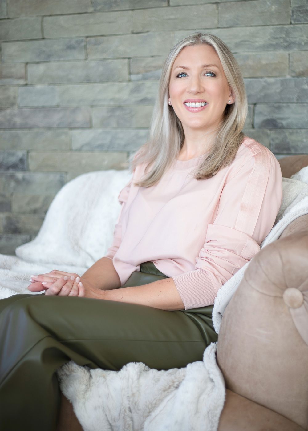 Person in pink sweater and olive pants relaxes on a beige couch with white pillows against a stone wall.