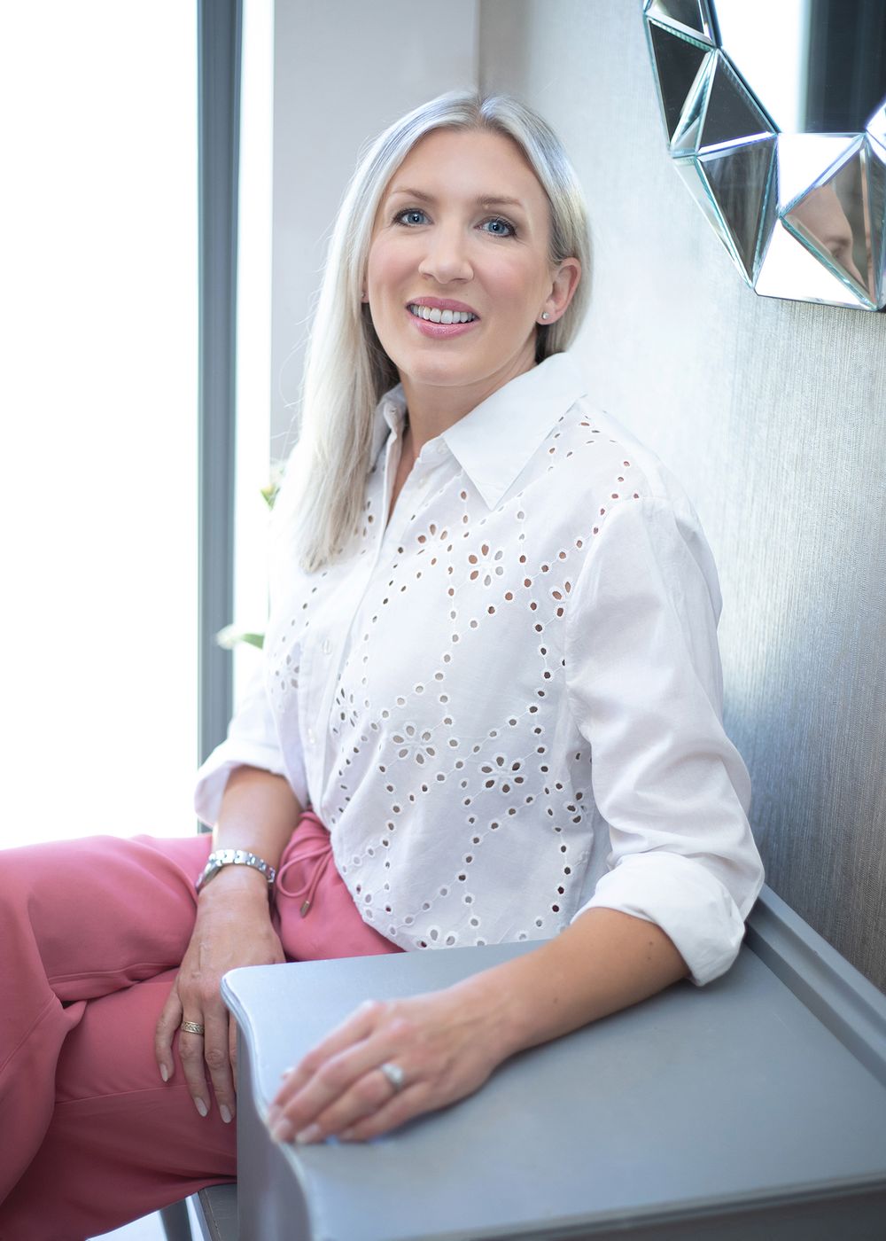 Woman in white eyelet blouse sits casually on gray bench against dark wall in professional setting.