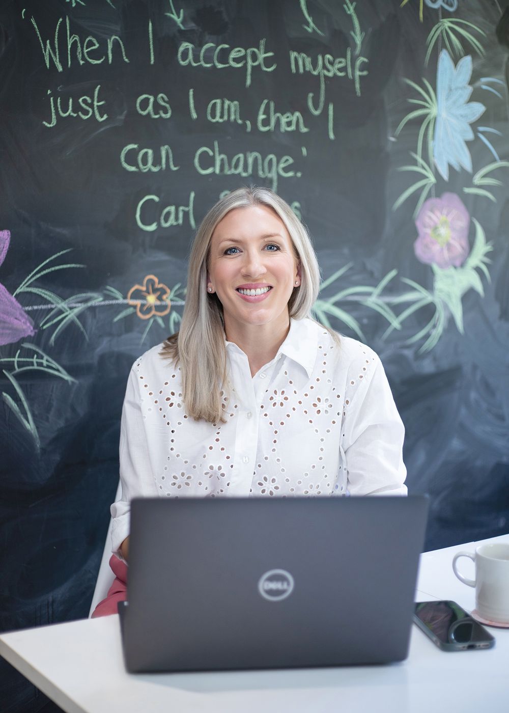 Someone in a white eyelet blouse works at a laptop against a chalkboard wall with colorful floral drawings.