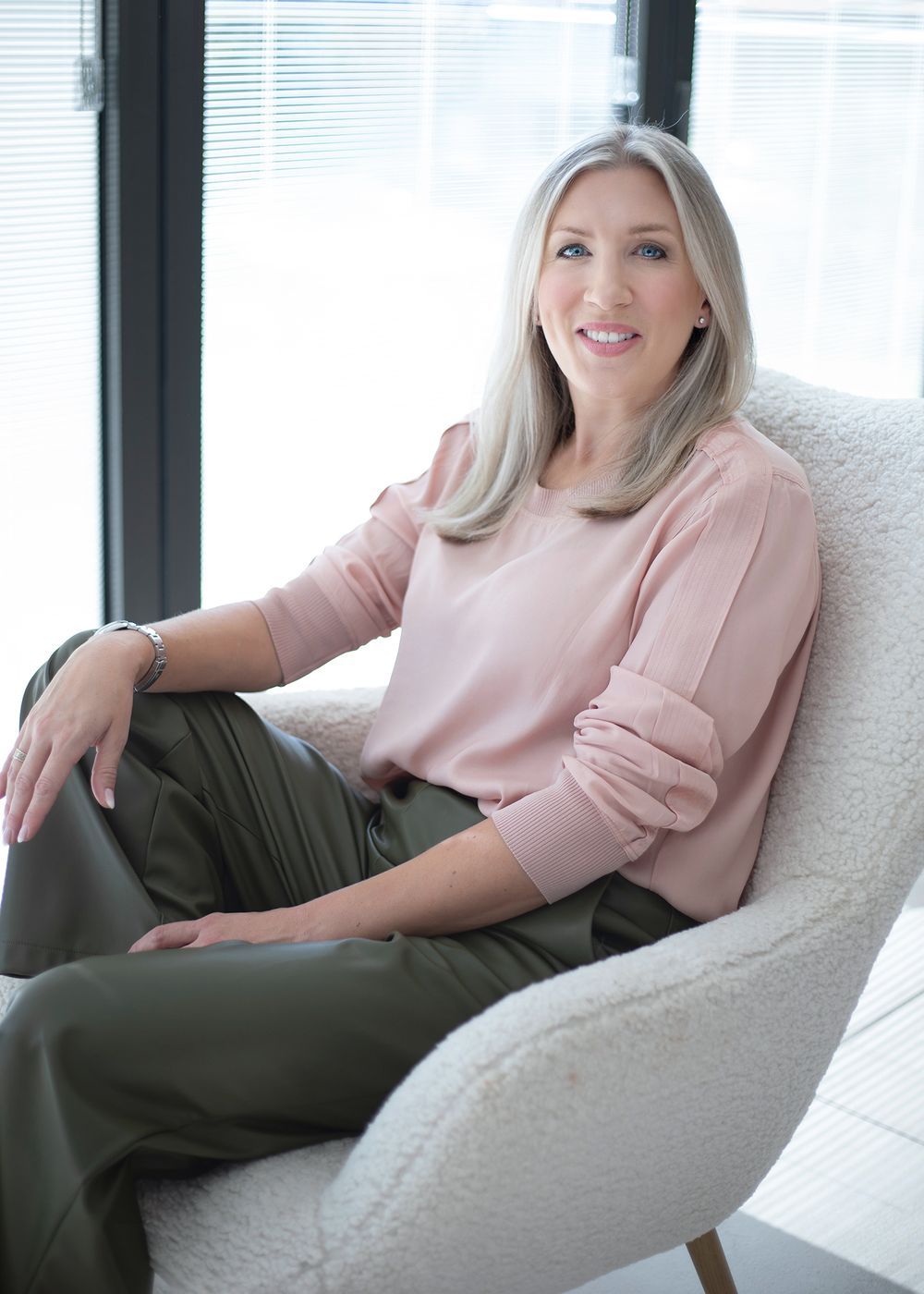 Woman in pink sweater and olive pants relaxes on white couch in bright modern room.