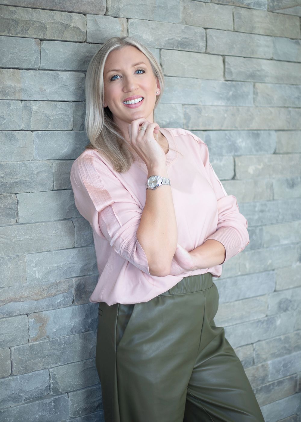 A person in a pink blouse and olive pants poses casually against a grey stone wall.