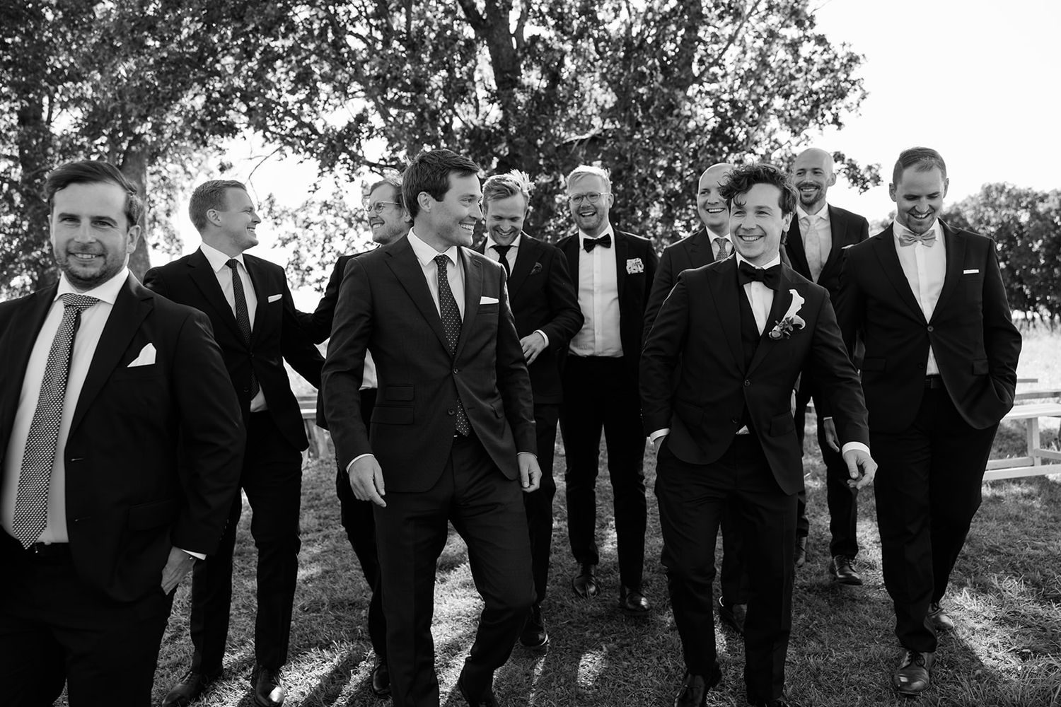 Groomsmen in black tuxedos walk in a line outdoors during a formal wedding photo session.