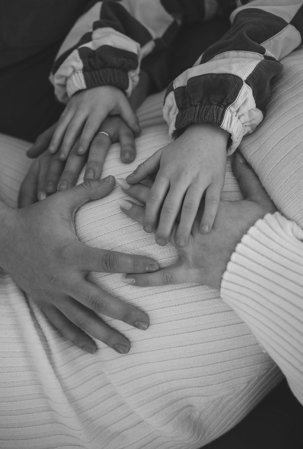 Black and white photo of family hands resting on a pregnant belly, forming a heart shape together.