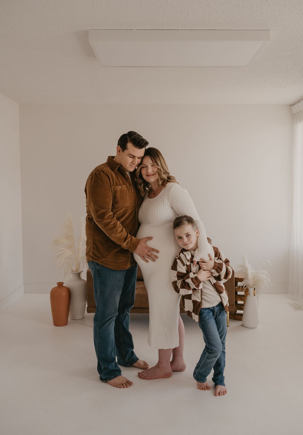 A couple and young child pose for a maternity photo in a bright white studio, the pregnant woman in a white ribbed dress.
