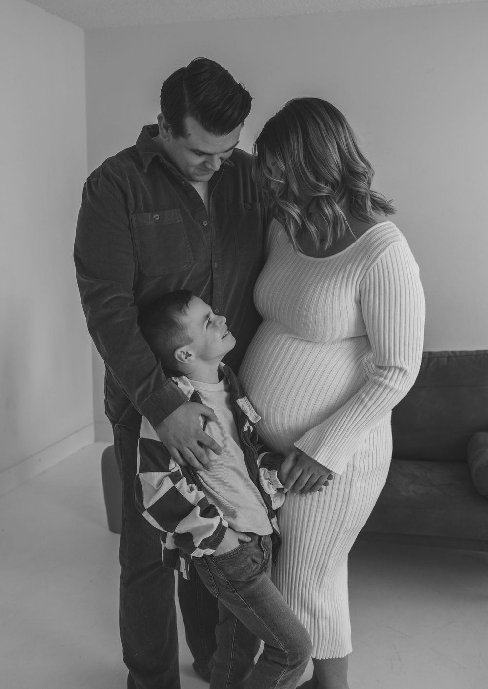 Black and white maternity photo of pregnant woman, man, and young boy looking at baby bump indoors.