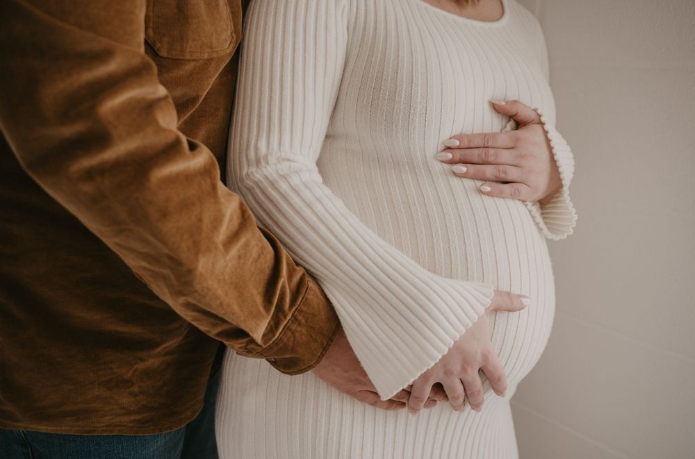 Pregnant woman in white ribbed dress cradling baby bump with partner in brown jacket beside her.