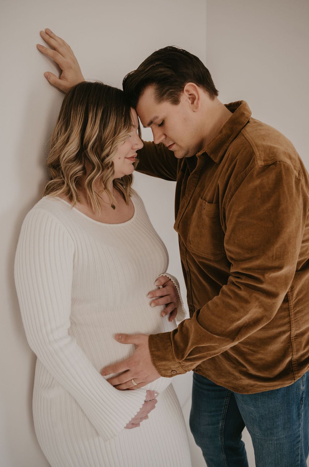 An intimate maternity photo of a couple touching foreheads, the man in a brown corduroy shirt cradling the pregnant belly.