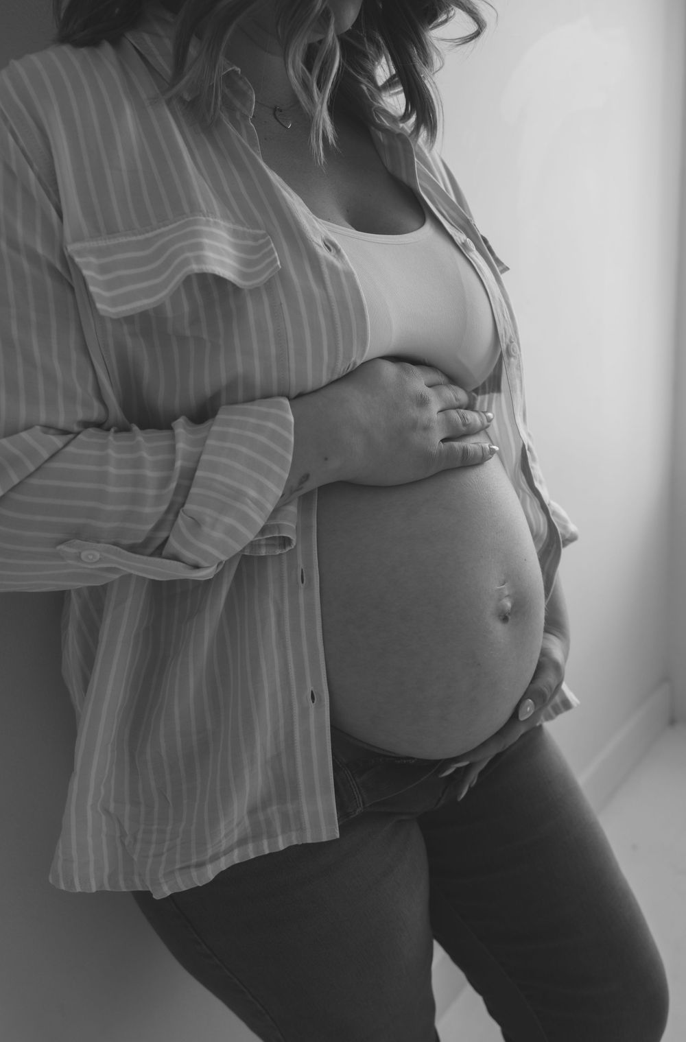Black and white close-up of a pregnant woman in an open striped shirt, gently holding her bare baby bump.