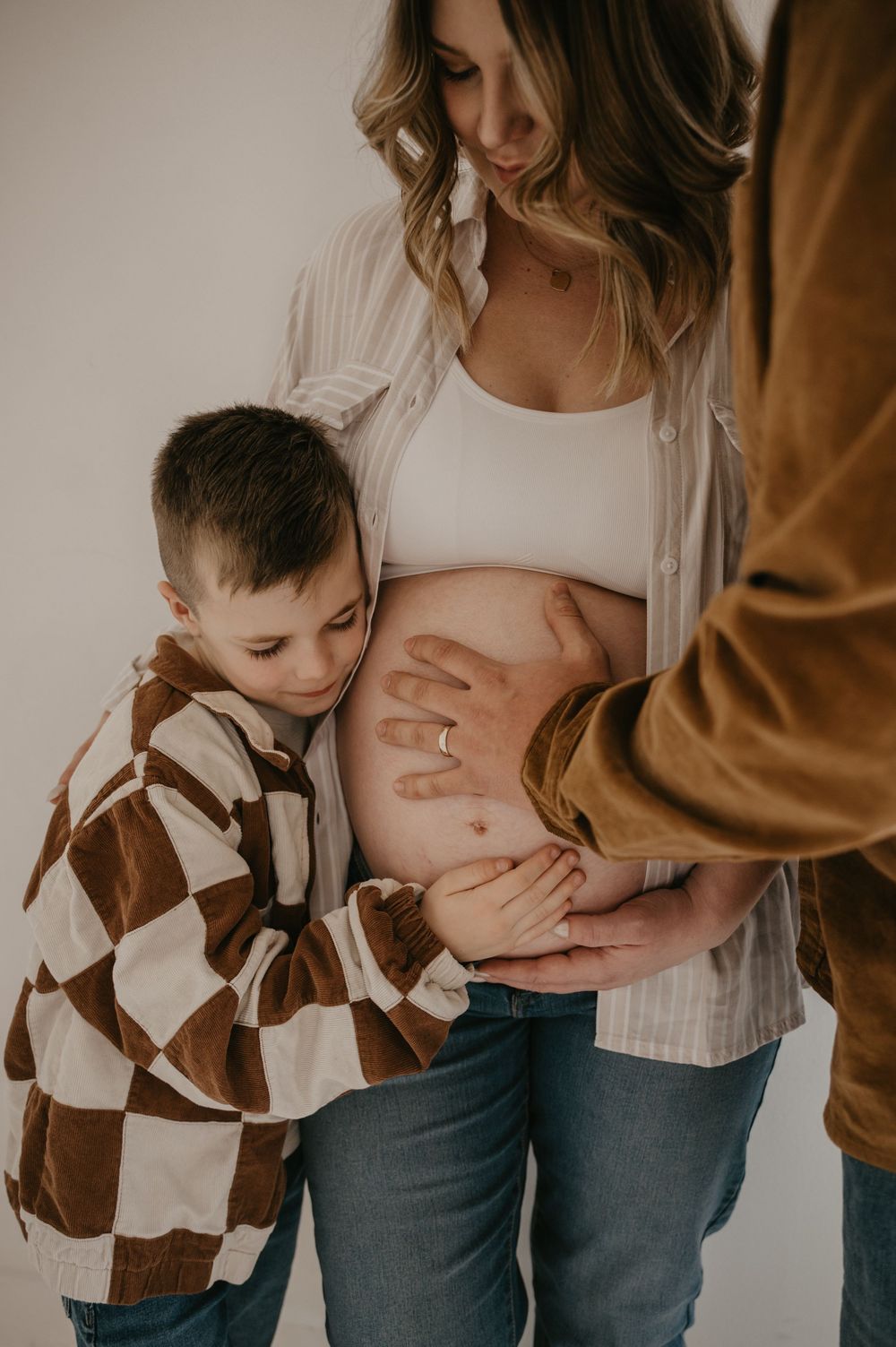 A young boy lovingly kisses a pregnant woman's baby bump while a man gently places his hand on her belly.