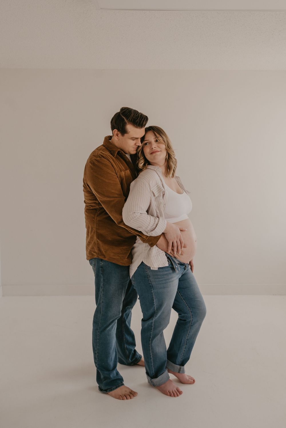 Couple sharing tender maternity moment, man in brown jacket embracing pregnant woman in white top and jeans.