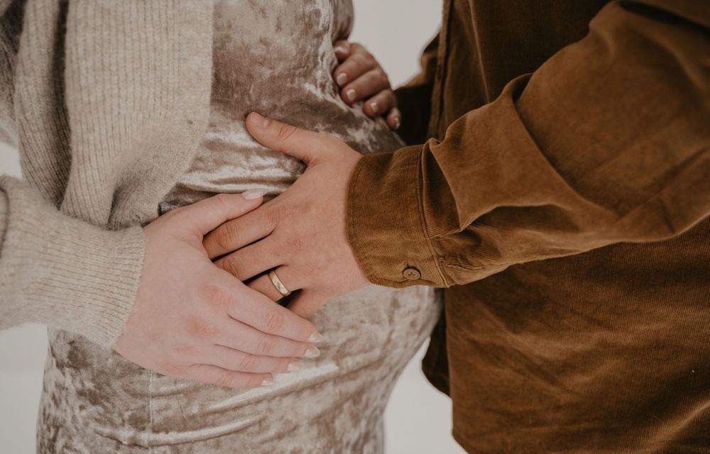 Couple tenderly holding pregnant belly, woman wearing ring, dressed in cozy neutral tones for maternity photo.