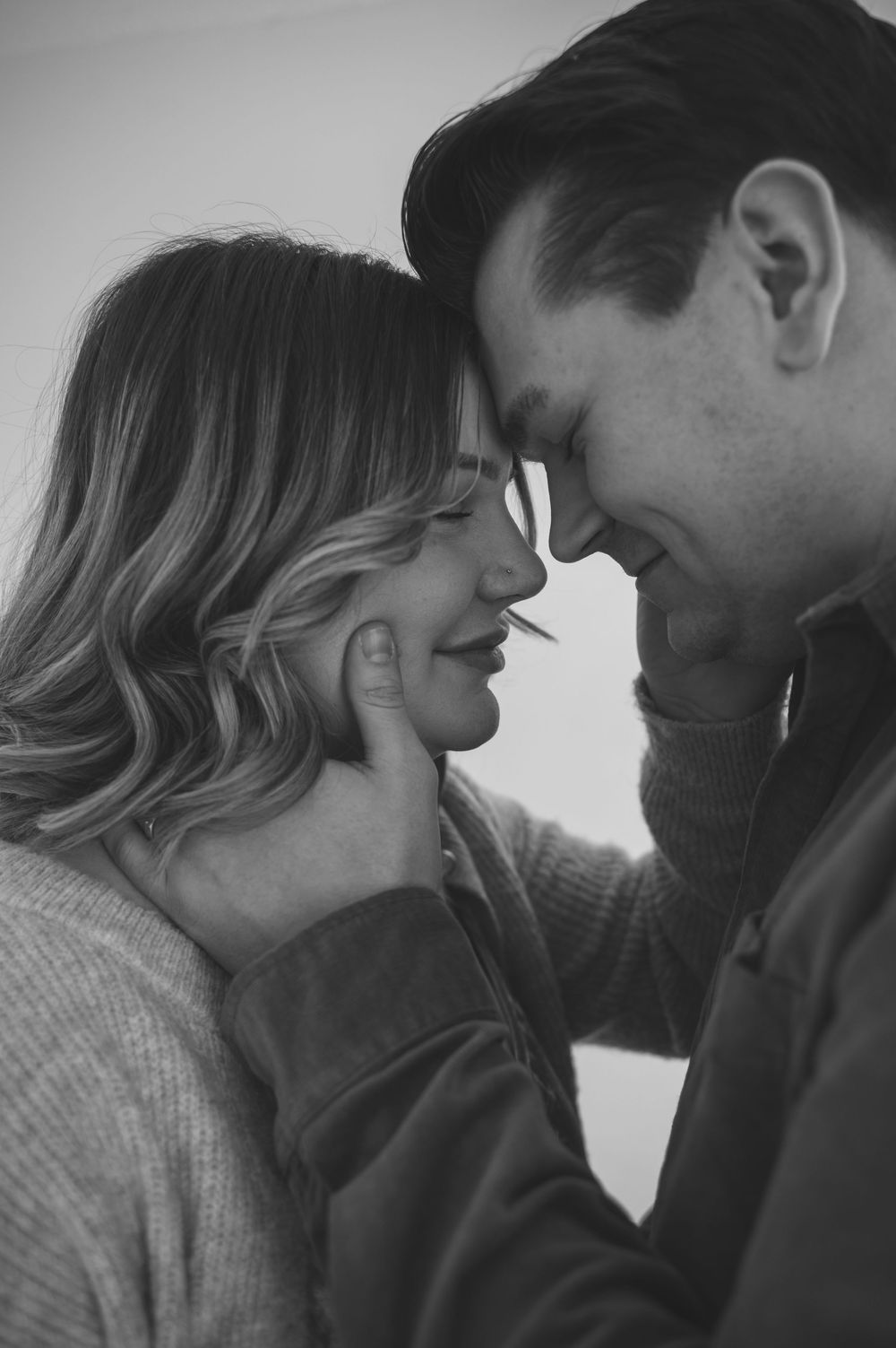 Black and white romantic photo of a smiling couple touching foreheads, the woman tenderly cupping the mans face.