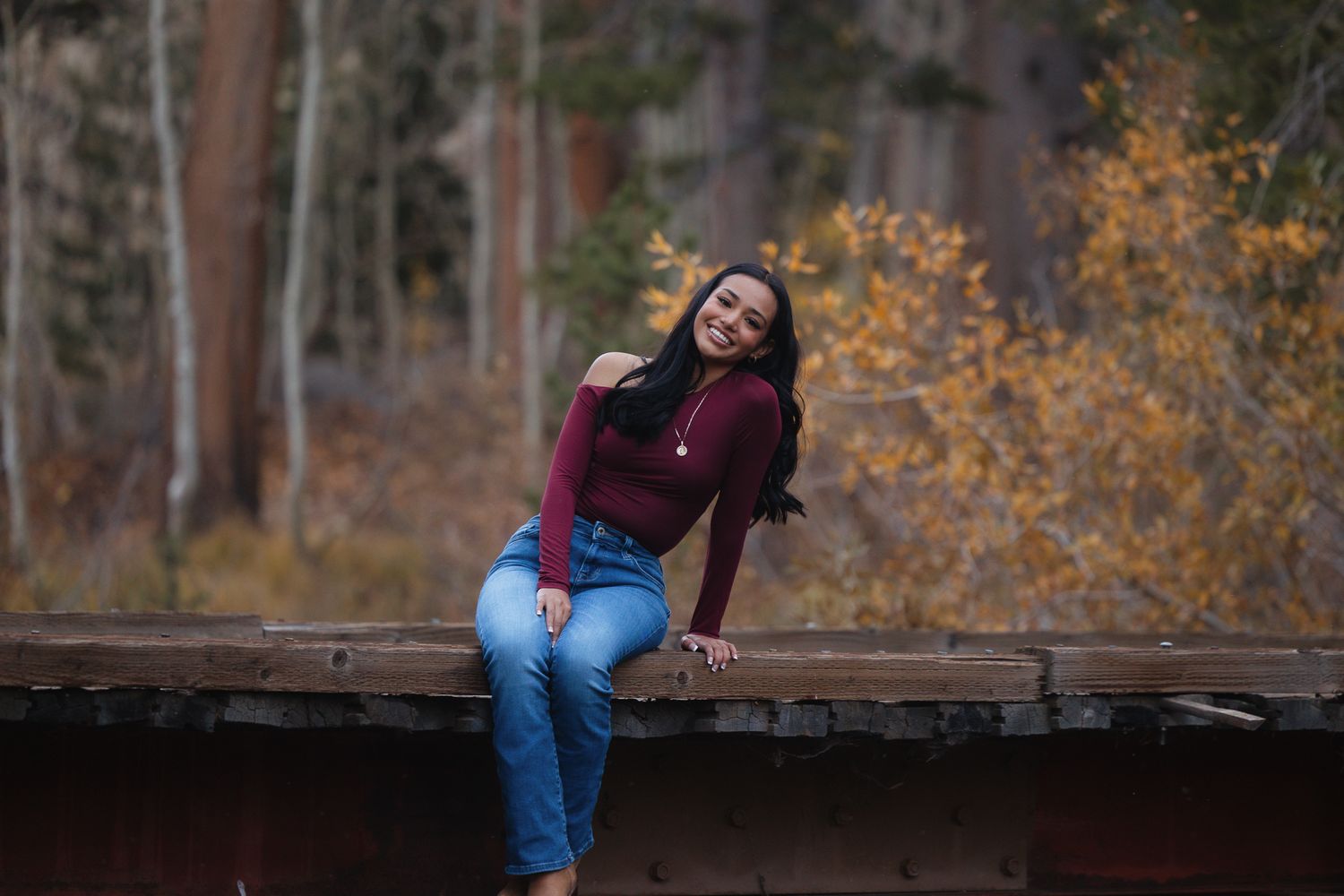 A person in a burgundy sweater and jeans sits casually on a wooden bridge with autumn foliage in the background.