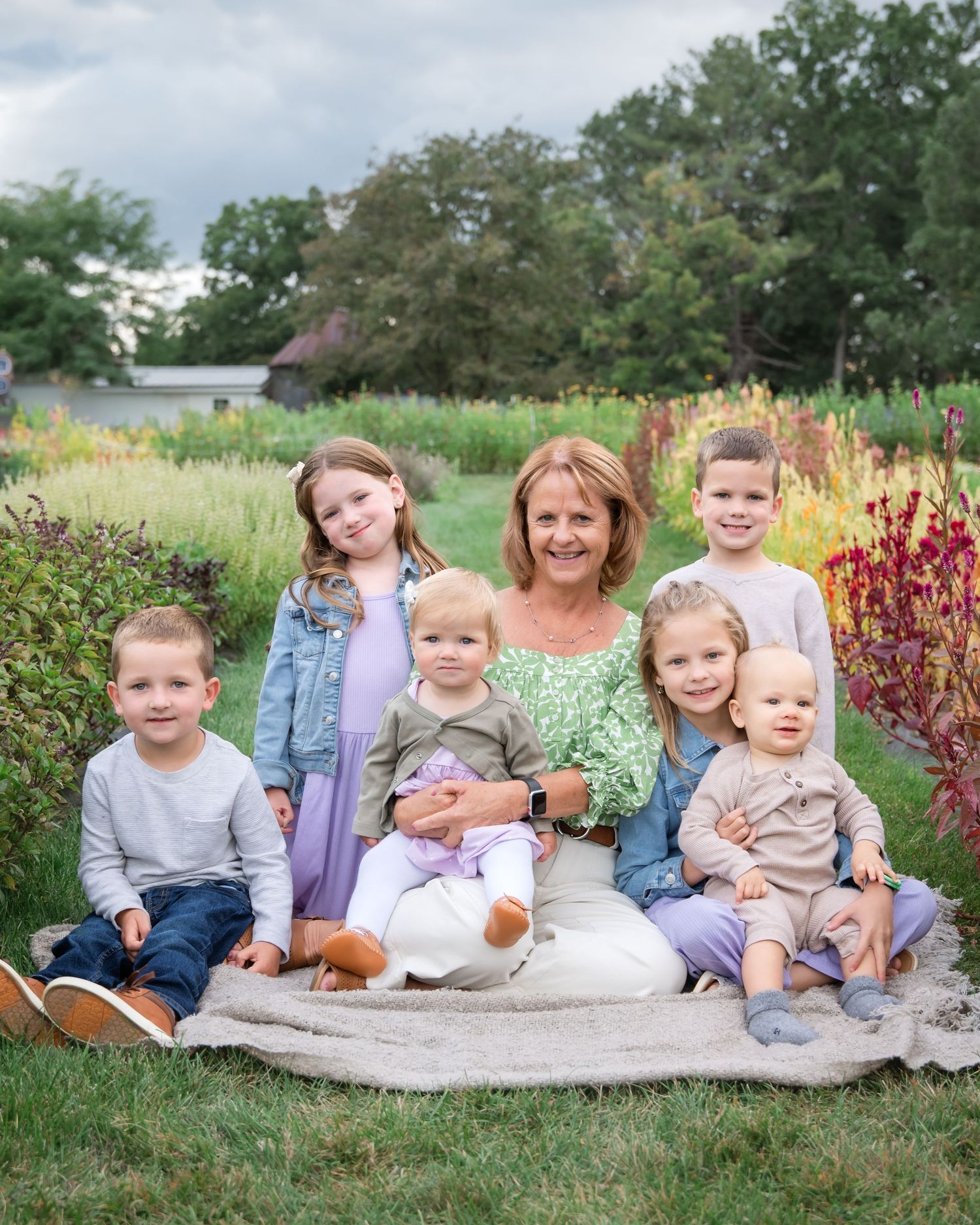 A family group sits together on a blanket in a garden with colorful flowers and greenery in the background.