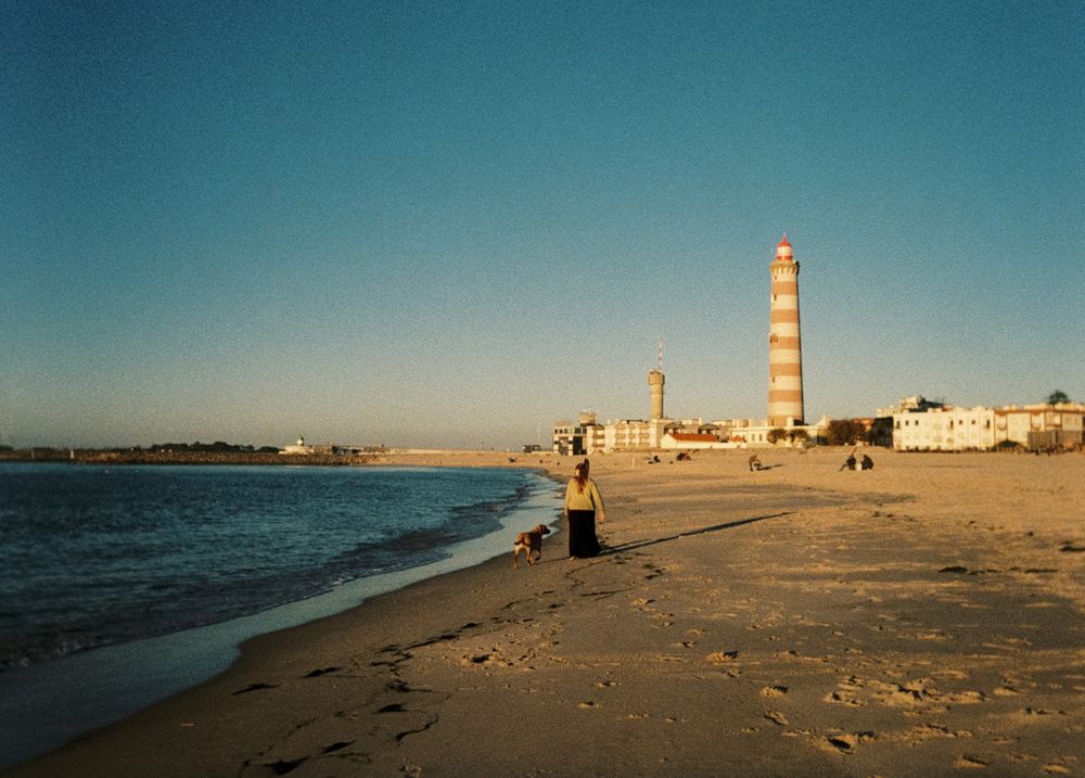 Sandy beach at sunset with a striped lighthouse and tower in the background near calm ocean waters.