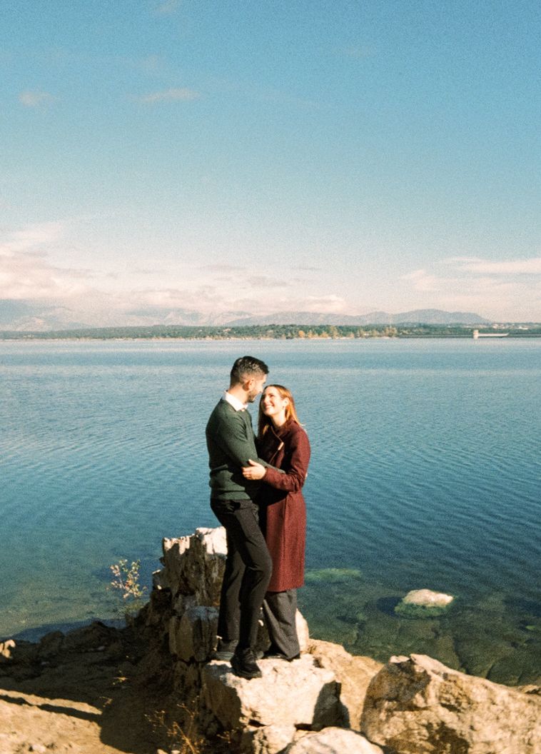 Couple embracing on rocky shoreline with blue lake and mountain backdrop on a sunny day.