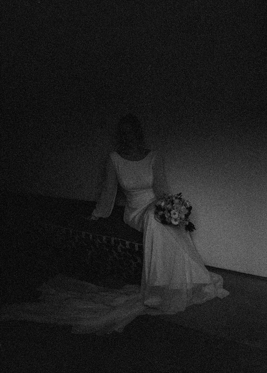 A bride in a white gown holds a bouquet while seated in a dimly lit, moody black and white photograph.